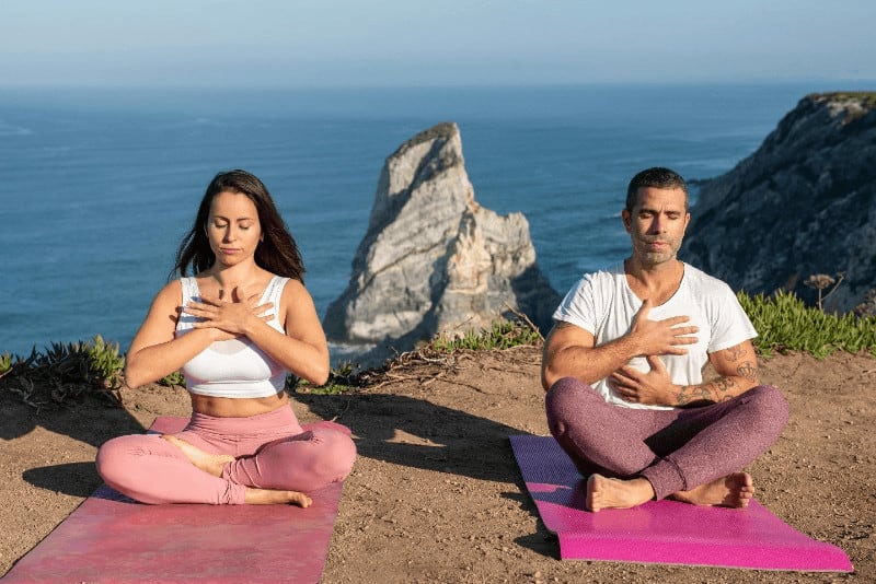 man and woman sitting on their yoga mats, practicing breathing techniques with a beautiful ocean view background