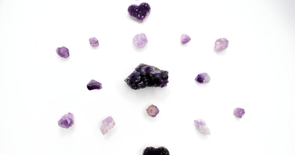 A collection of purple crystals, varying in size and shape, arranged on a white background. The central crystal is significantly larger and darker than the others, which are lighter in color and mostly have a hexagonal shape.