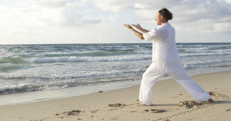 The image shows a person standing on a sandy beach near the ocean, performing what appears to be a Tai Chi or similar meditative exercise. The person is dressed in white, loose-fitting clothing and is in a stance with one leg bent forward and arms extended outward. The ocean waves are gently crashing onto the shore, and the sky is partly cloudy. This image is interesting and relevant as it captures a serene and peaceful moment, highlighting the practice of mindfulness and physical exercise in a natural, calming environment. There are no math or homework problems in the image.