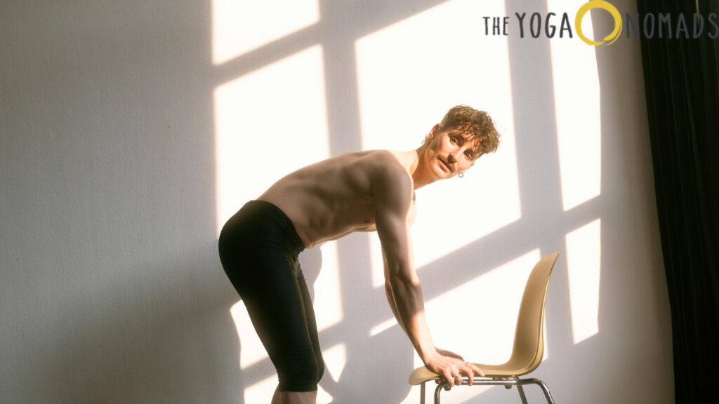 The image shows a person performing a yoga pose using a chair. The person is shirtless and wearing black shorts, bending forward with hands resting on the back of a chair. The background features a wall with shadows cast by a window, and the text "THE YOGA NOMADS" is visible in the upper right corner. This image is relevant as it demonstrates a yoga pose that can be adapted for chair yoga, which is beneficial for people with limited mobility.