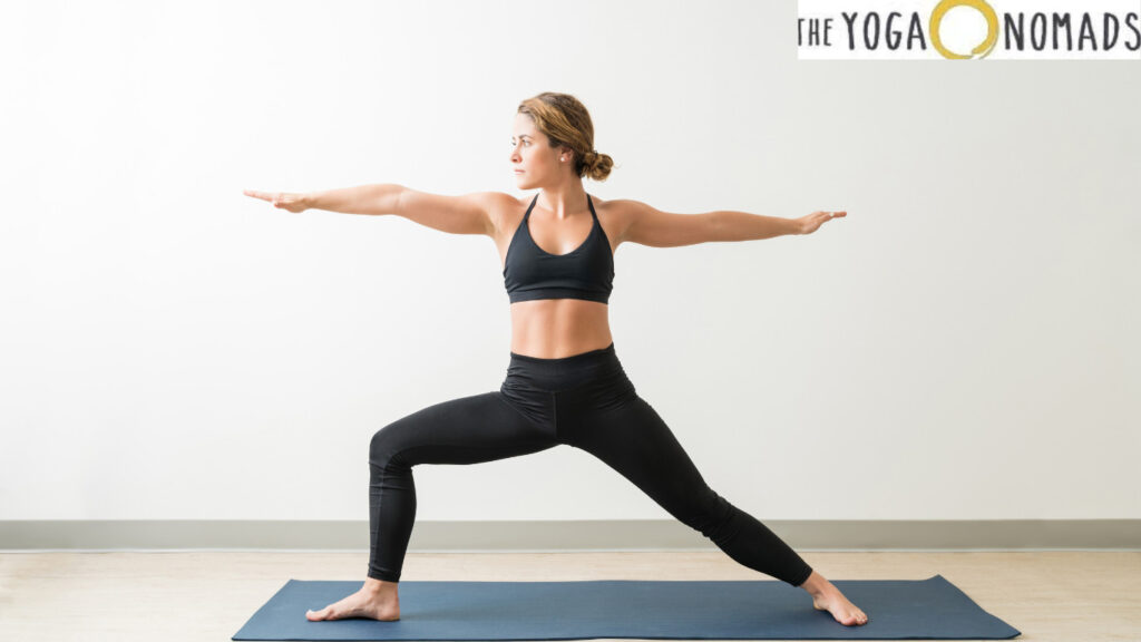 A person is performing a yoga pose on a blue yoga mat. The individual is dressed in black athletic wear, consisting of a sports bra and leggings. The pose involves standing with legs apart, one foot pointing forward and the other foot pointing sideways, with arms extended horizontally. The background is a plain white wall.