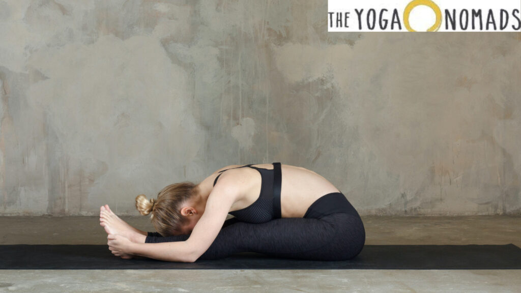 A person is performing a seated forward bend yoga pose on a black yoga mat. The person is sitting with legs extended straight in front, bending forward to touch the feet with the hands, and resting the head on the knees. The background is a plain, textured wall.