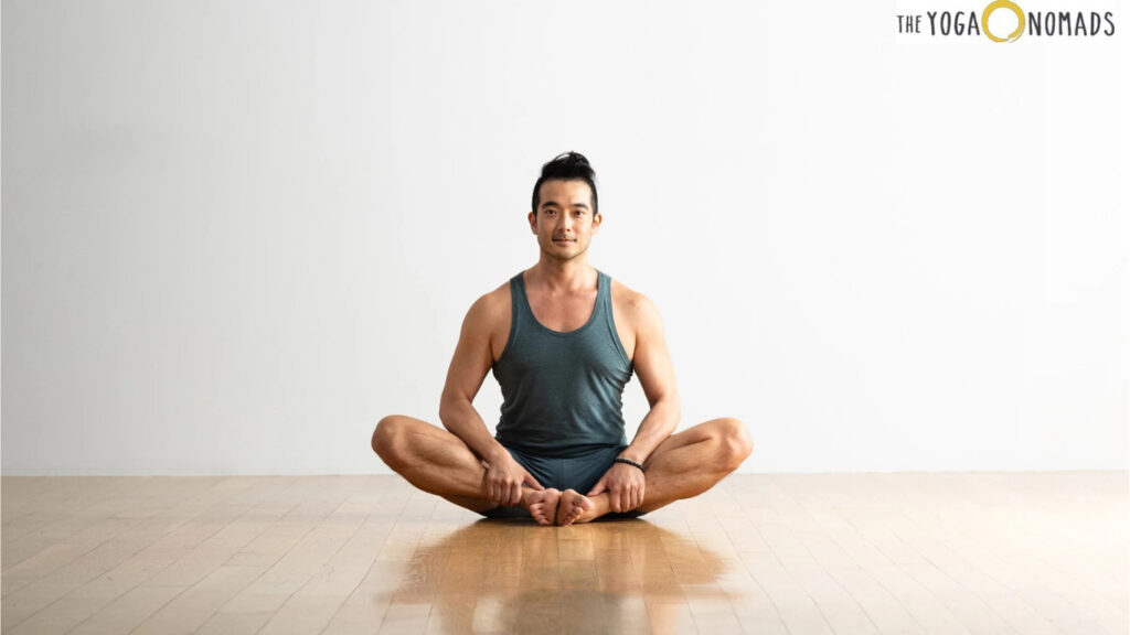 The image shows a person sitting on a wooden floor in a yoga pose known as the Butterfly Pose or Baddha Konasana. The person is wearing a dark tank top and shorts, with hands holding the feet together and knees bent outward. The background is plain white, and the logo "The Yoga Nomads" is visible in the top right corner. This image is relevant as it demonstrates a common yoga pose that helps in stretching the inner thighs, groin, and knees, promoting flexibility and relaxation.