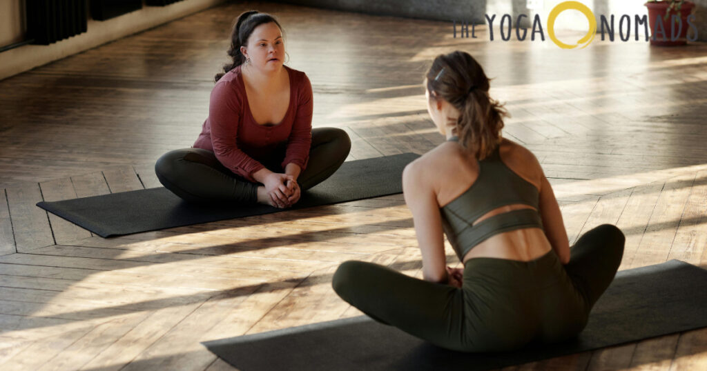 The image shows two people performing the butterfly stretch on yoga mats in a well-lit room with wooden flooring. The individual on the left is wearing a red long-sleeve shirt and dark pants, while the person on the right is dressed in a green sports bra and matching leggings. Both are seated with the soles of their feet pressed together and knees bent outward, showcasing the butterfly stretch, which is beneficial for improving hip flexibility and easing tension in the groin area.