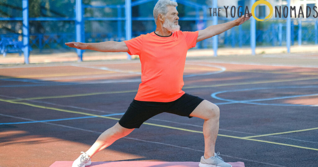 The image shows a person performing the Warrior 2 pose on a yoga mat outdoors on a sports court. The individual is standing with one leg bent at a 90-degree angle and the other leg extended straight behind. Their arms are stretched horizontally, parallel to the ground, creating a strong and balanced posture. The person is wearing a bright orange shirt, black shorts, and white sneakers. The background includes blue fencing and a blurred outdoor setting, emphasizing the energetic and athletic vibe of the pose.